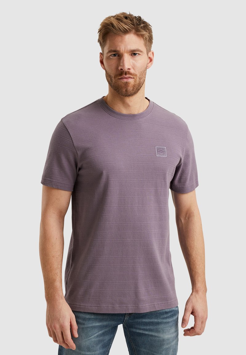 Camiseta de jersey morada de manga corta con un patrón texturizado, cuello redondo y un pequeño parche de logo en el pecho. Combinada con jeans azules.