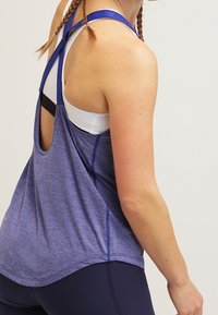 Fioletowy sportowy top na ramiączkach z wycięciem na plecach i kontrastującym przeszyciem, noszony na białym sports-bra.