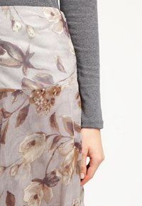 Höyhenenkevyt, kukkakuvioinen vaate puoliläpinäkyvästä, kevyestä materiaalista, jossa on beigejä ja ruskeita sävyjä. Harmaa pitkähihainen paita näkyvissä.