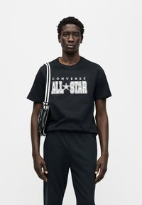 Μαύρο βαμβακερό t-shirt με το κείμενο "CONVERSE ALL-STAR" σε λευκό, στενή εφαρμογή, σε συνδυασμό με μαύρο παντελόνι και μαύρη τσάντα με λευκές λωρίδες.