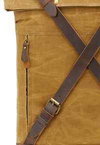 Brun canvasryggsäck med läderremmar, en dragkedjeficka och mässingsdetaljer. Texturerad yta med en robust design.
