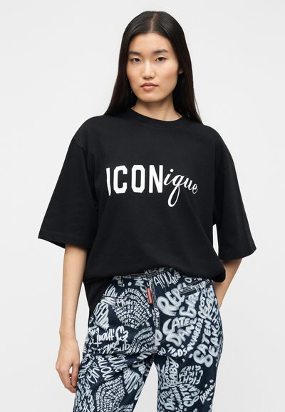 Μαύρο υπερμεγέθες μπλουζάκι με λευκό γραφικό κείμενο «ICONique». Συνδυάζεται με μπλε τζιν με λευκά γραπτά μοτίβα, παρουσιάζοντας έναν χαλαρό σχεδιασμό.