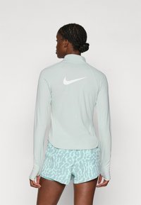 Světle zelené tričko s dlouhým rukávem na sport s vysokým límcem a bílým logem Nike na zádech, spárované s šortkami s mentolovým vzorem.
