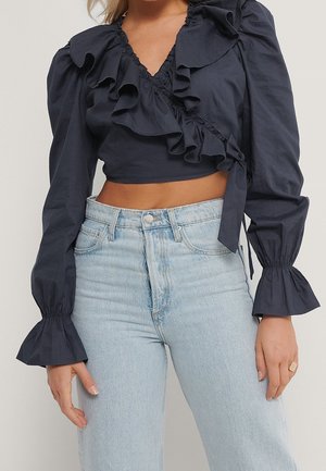 Blusa cropped blu navy con colletto arricciato, maniche lunghe e polsini svasati. Abbinata a jeans chiari a vita alta. Tessuto in cotone.