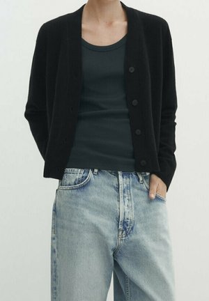 Person trägt einen schwarzen, geknöpften Cardigan, ein dunkelgrünes ärmelloses Top und hellblaue, hoch taillierte Jeans mit einer Hand in der Tasche.