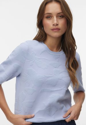 Woman with long wavy brown hair wearing a light blue short-sleeve sweater with a subtle floral pattern and hoop earrings.