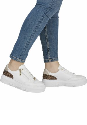 Bílé tenisky s nízkým střihem s leopardími vzory na patě a zlatým zipem, nošené s modrými úzkými džíny na zkřížených nohách.