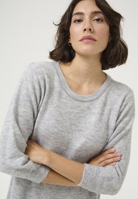 Pull en tricot gris avec un col rond côtelé, des manches trois-quarts et une texture douce, comprenant des embellissements subtils au niveau du col.