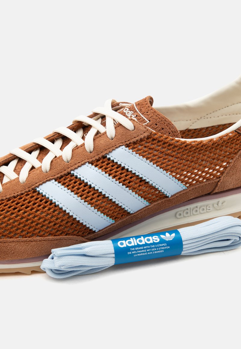 Baskets Adidas en mesh marron avec des rayures bleu clair et des lacets blancs, placées à côté d'une paire de lacets Adidas bleu clair dans un emballage de marque.