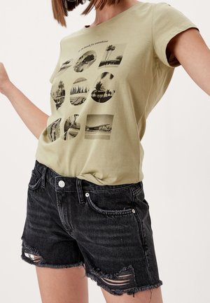 Kvinde iført beige grafisk t-shirt med sort-hvide naturbilleder og sorte frynset denimshorts foran en ensfarvet baggrund.