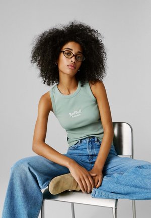 Mintunvihreä ribbineulottu hihaton toppi, jossa teksti "RedBull Racing", yhdistetty vaaleansinisiin korkeavyötäröisiin farkkuihin ja beigen värisiin nilkkureihin, istumassa tuolilla.