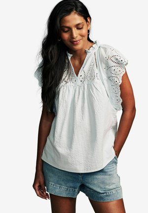 Camicia bianca in cotone con dettagli a occhiello, corpettino arricciato e maniche a volant, abbinata a pantaloni corti in denim. Tessuto semplice e strutturato.
