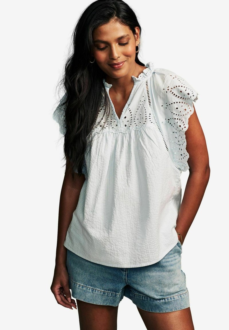 Camicia bianca in cotone con dettagli a occhiello, corpettino arricciato e maniche a volant, abbinata a pantaloni corti in denim. Tessuto semplice e strutturato.