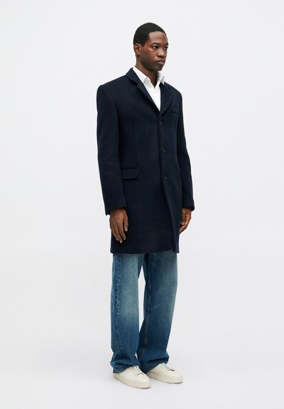 Navy wool blend coat with tailored lapels, single-breasted design, and side pockets, paired with light blue denim jeans and white sneakers.