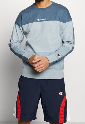 Felpa Champion blu e grigia con maniche lunghe, scollo a girocollo e logo; abbinata a pantaloncini blu navy con dettagli laterali rossi e bianchi.