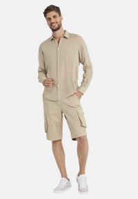 Camicetta beige in lino a maniche lunghe con bottoni, abbinata a pantaloni cargo beige con tasche laterali e indossata con sneakers bianche.
