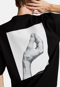 Camiseta de algodón negra con un gran gráfico de una mano en un gesto, contorneada en blanco en la parte posterior, con letras en negrita debajo.