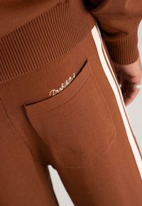 Pantaloni tricotati maro cu dungi laterale albe și un buzunar posterior brodat cu cuvântul "Interzis", purtați de o persoană cu inele.