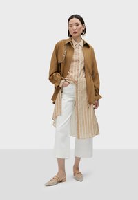 Brun jacka med dragsko, lager på lager över en beige och vit randig skjorta, tillsammans med vita vida byxor och beige spetsiga ballerina.