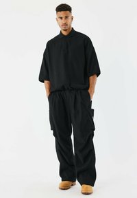 Polo shirt nera oversize con colletto ripiegato, abbinata a pantaloni cargo neri a vestibilità ampia con tasche laterali. L'outfit è completato da zoccoli color sabbia.