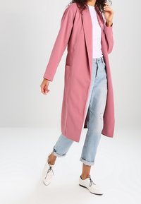 Lång rosa kappa med en slank design, med fickor och öppen front, kombinerad med ljusblå slitna jeans och vita skor.