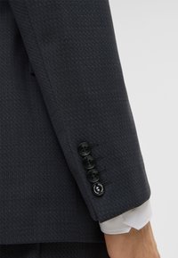Rękaw ciemnej, dopasowanej marynarki z teksturowanej tkaniny. Pięć czarnych guzików z dwoma dekoracyjnymi elementami. Widoczny mankiet białej koszuli.