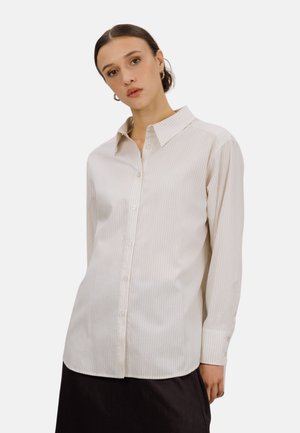 Kvinne med mørkt hår bundet bakover, iført en langermet beige stripet skjorte og svart skjørt, stående foran en hvit bakgrunn.