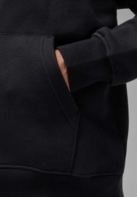 Felpa nera con tasca a marsupio, polsini e orlo a coste. Il materiale sembra morbido con una texture liscia. Vista ravvicinata della cucitura laterale.