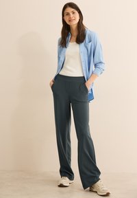 Jasnoniebieska koszula zapinana na guziki noszona na białym podkoszulku, w zestawie z ciemnozielonymi spodniami o szerokich nogawkach i beżowymi sneakersami. Gładki materiał z subtelnymi kieszeniami.