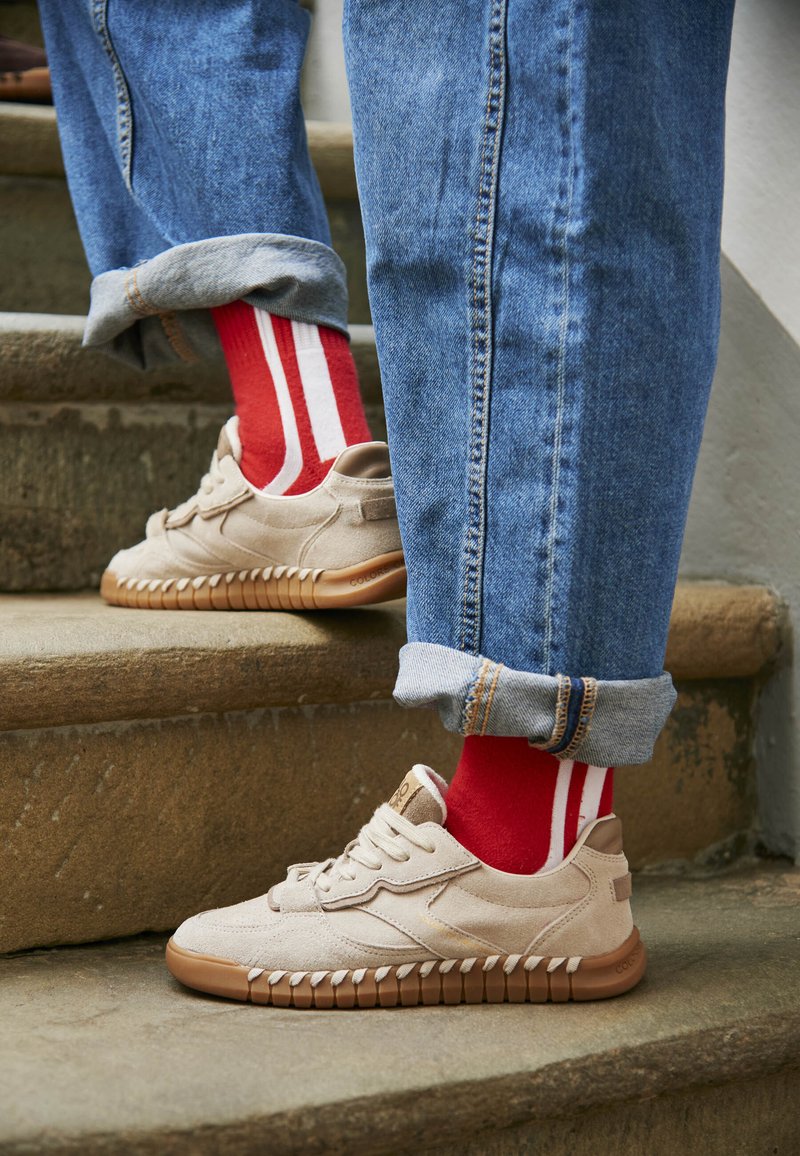 Helle beige Wildleder-Sneaker mit brauner Gummisohle, kombiniert mit hochgekrempelten blauen Jeans und roten Socken mit weißen Streifen.