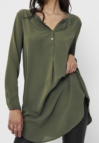 Blouse à manches longues vert olive avec patte de boutonnage, ourlet arrondi et tissu texturé. Présente un léger drapé et une finition douce.