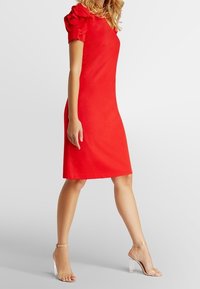 Robe rouge avec manches ballons, longueur genoux, silhouette ajustée. Tissu lisse, associée à des sandales à talons transparents. Design simple sans motifs visibles.