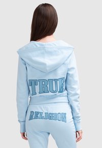 Jasnoniebieska bluza z kapturem z dużym haftowanym napisem "TRUE" na plecach. Teksturowany materiał i skrócony fason. Pasujące spodnie dresowe z akcentem "RELIGION".