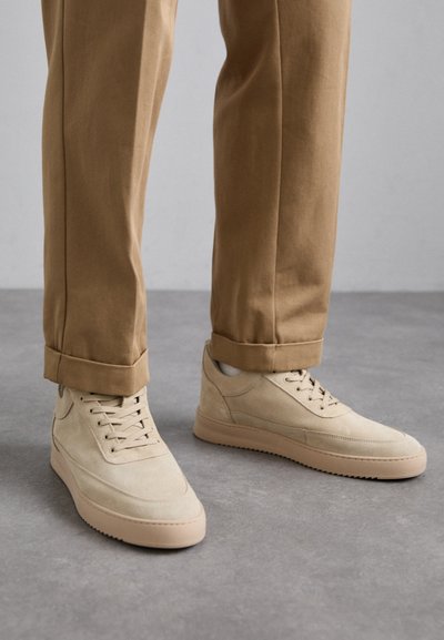 Sneakers di suede beige con una texture liscia, design con lacci e suole in gomma, abbinati a pantaloni tan arrotolati.