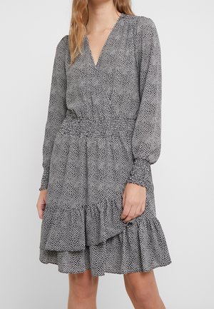 Kobieta ubrana w czarno-białą sukienkę kopertową w groszki z długimi rękawami, elastyczną talią, marszczonymi mankietami i falbaną u dołu sięgającą powyżej kolan.