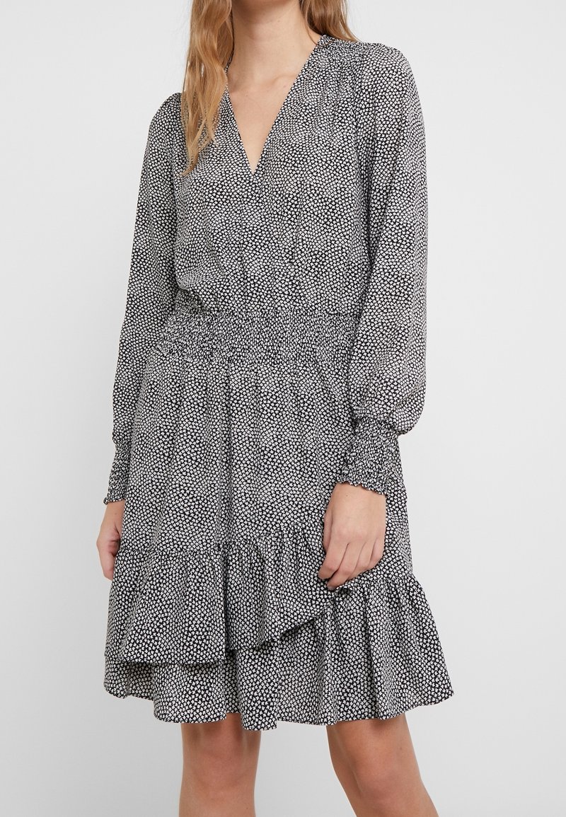 Femme portant une robe portefeuille à pois noirs et blancs avec manches longues, taille élastique, poignets smockés et ourlet volanté au-dessus des genoux.