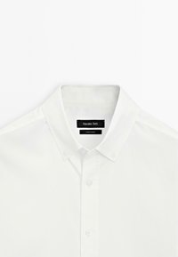 Chemise blanche à boutons avec un col boutonné, étiquette de marque visible "Massimo Dutti" et étiquette "Fabriqué en Turquie" à l'intérieur du col.