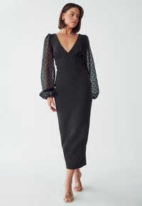 Čierne midi šaty s hlbokým výstrihom do V, priliehavým korzetom a priesvitnými balónovými rukávmi, ktoré sú zdobené čiernymi bodkami. Hladká tkanina s elegantným strihom.