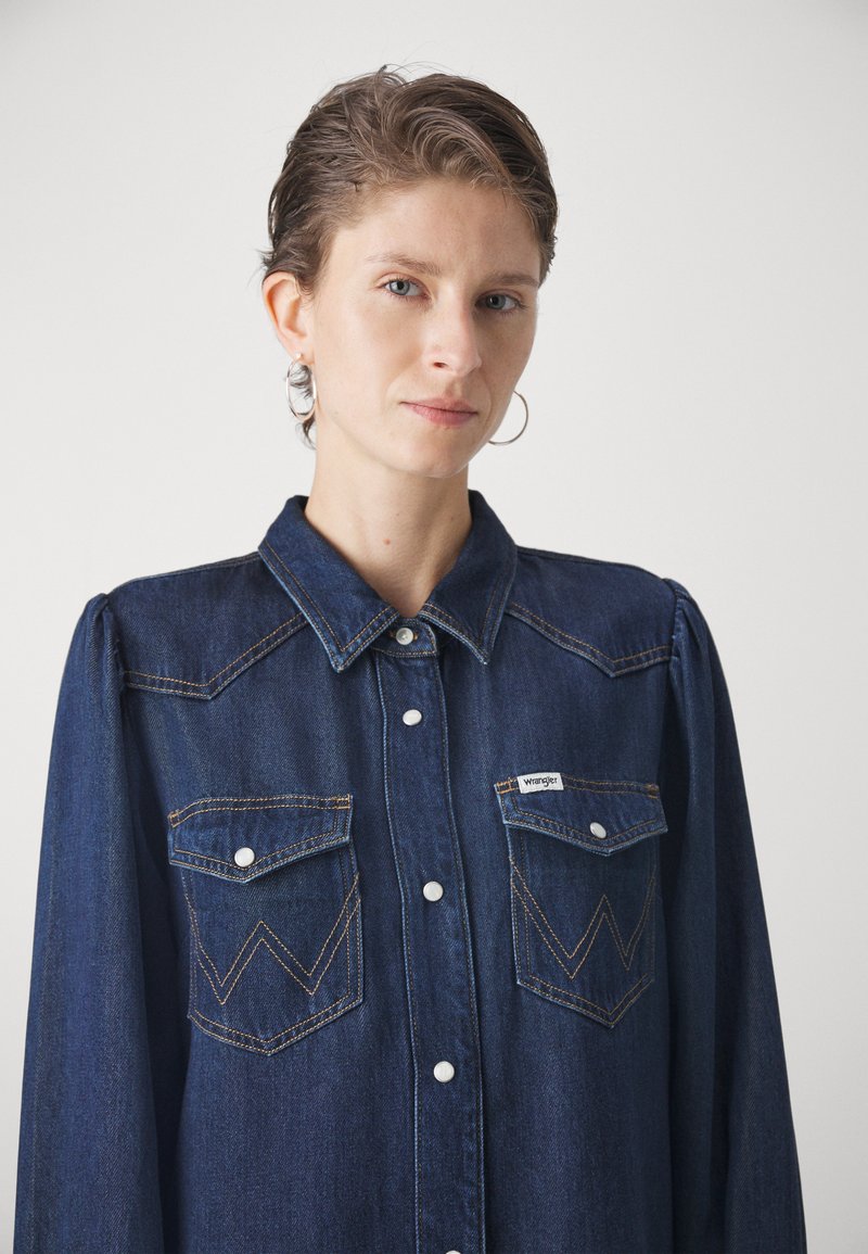 Chemise en denim de couleur bleu foncé avec un devant boutonné, deux poches poitrine ornées de coutures en zigzag, et un col. Étiquette Wrangler visible.