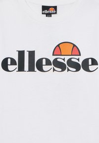 Fehér pamut póló, amelyen egy nagy fekete "ellesse" márkajanló látható, felette egy narancssárga és vörös félkörrel, ami sportos dizájnra utal.