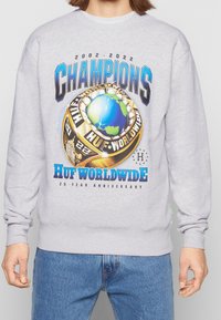 Felpa grigia con una grande grafica di un anello di campionato con un globo, il testo "CHAMPIONS" e "HUF WORLDWIDE 20 YEAR ANNIVERSARY."