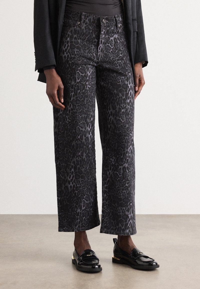 Personne portant un pantalon large imprimé léopard noir et gris, des mocassins noirs et une veste noire, visible de la taille aux pieds.