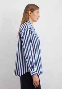 Chemise à rayures bleues et blanches avec un col, des manches longues et des poignets à boutons. Tissu lisse avec une coupe décontractée et un ourlet arrière plus long.
