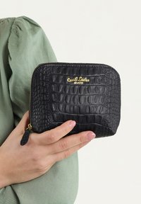 Melnā krokodila ādā iešūta somiņa ar zelta logo, ar rāvējslēdzēju un teksturētu virsmu. Turēta rokā pret zaļu fona.