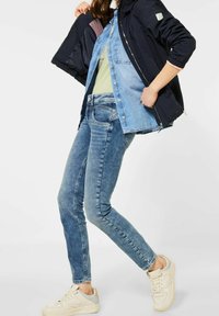 Marinblå bomberjacka, ljusblå denimskjorta, ljusgrön topp och skinny blå jeans med en blekt tvätt; krämfärgade sneakers med grå accenter.