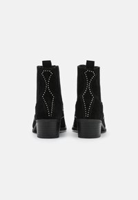 Botines de ante negro con un tacón bloque de altura media, que presentan tachuelas plateadas decorativas y costuras curvas en la parte trasera.