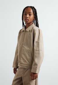 Beige zip-up jacket with a collared neckline, featuring a left chest pocket and ribbed cuffs, paired with matching trousers. Smooth texture.