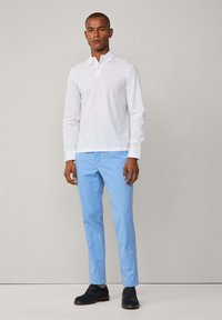 Weißes langärmliges Polohemd mit Kragen, kombiniert mit hellblauen Hosen. Die Schuhe sind dunkelblau. Glattes Gewebe, taillierte Passform, keine Muster.