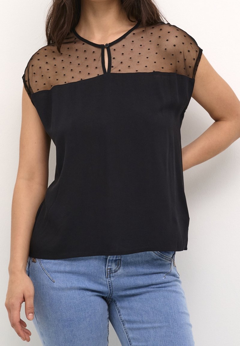 Haut noir avec tissu transparent à pois aux épaules, doté d'un col rond et de manches courtes, associé à un jean bleu clair.