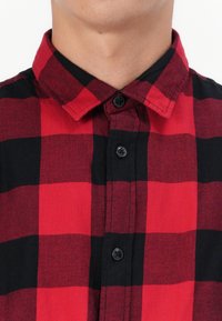 Primer plano de una persona que lleva una camisa de cuadros roja y negra con el botón superior abrochado.